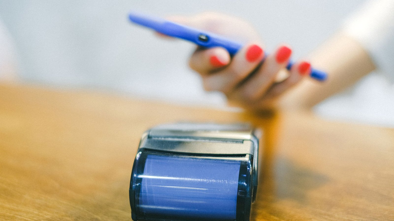 A hand holding a smartphone over a payment terminal on a wooden counter to make a contactless payment.