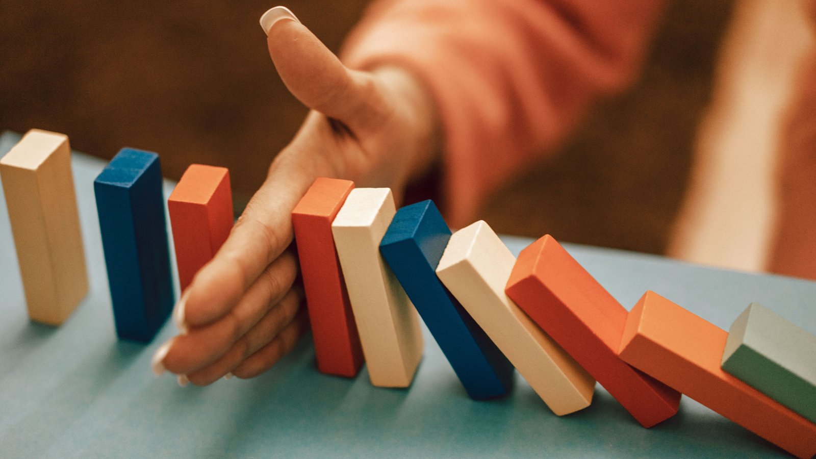 A hand stops a row of falling blue, orange and white wooden blocks on a table, like a line of dominoes.