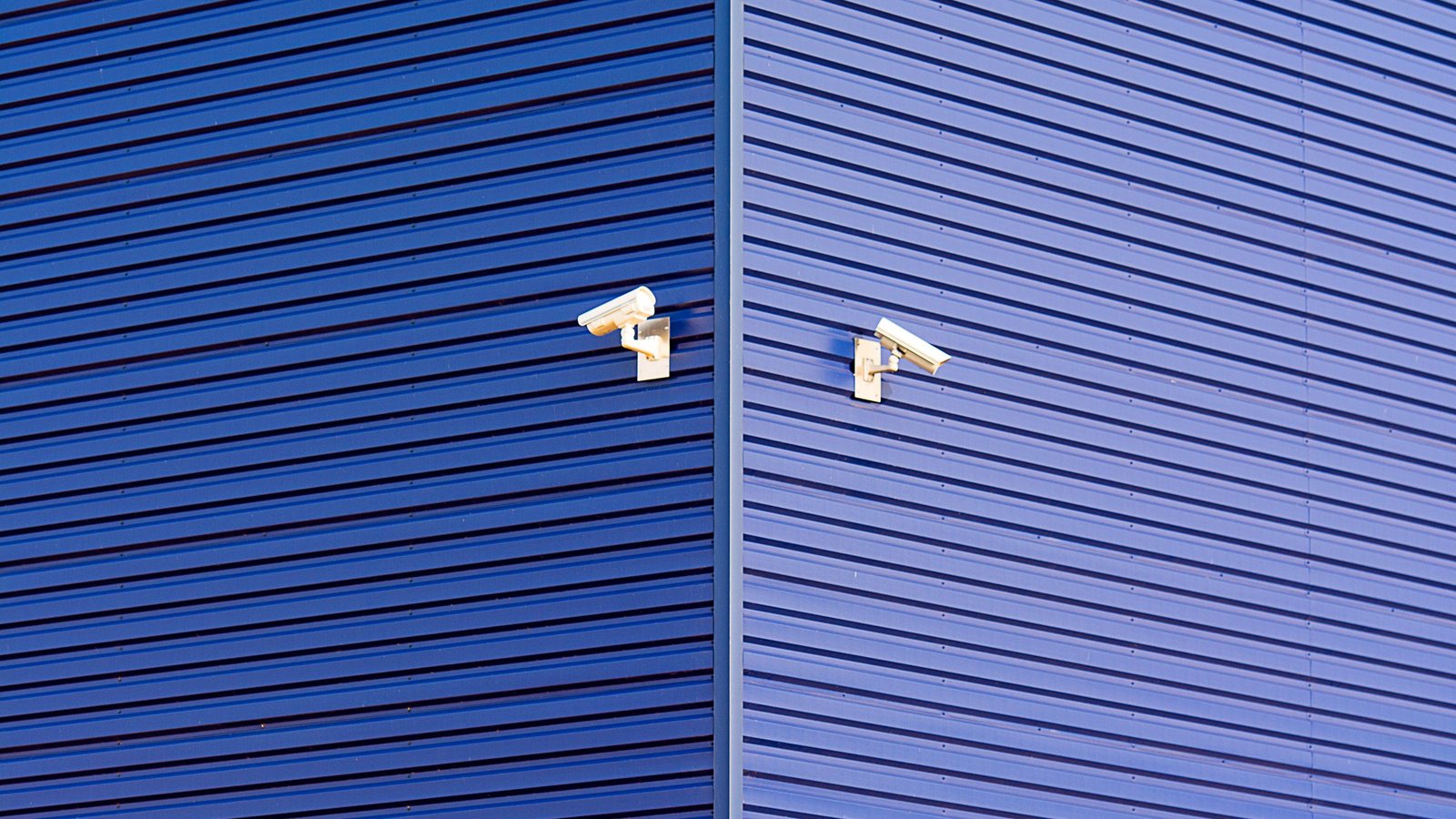 Two white CCTV cameras mounted on the corner of a blue ribbed metal building wall