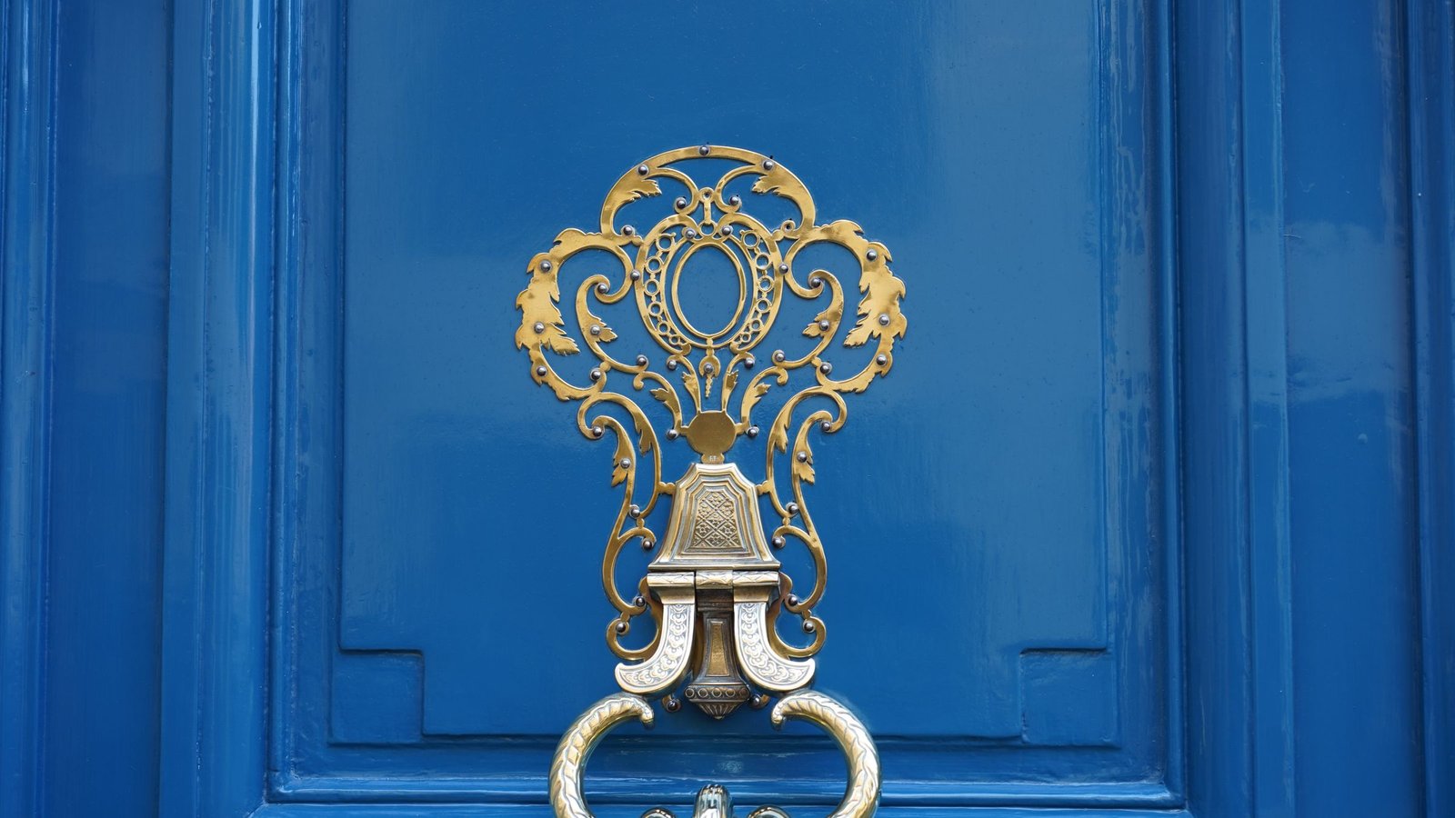 Close-up of an ornate polished brass door knocker mounted on a glossy blue wooden door panel, with no text or logos visible.